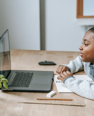Uczeń szkoły podstawowej siedzi przy biurku i uczestniczy w zajęciach online na laptopie, obok leży zeszyt do notatek, ołówek i linijka. Chłopiec skupia się na ekranie podczas lekcji, co dobrze oddaje ideę kursów dla ósmoklasistów online Symposio przygotowujących do egzaminu ósmoklasisty – więcej informacji można znaleźć na stronie organizatora